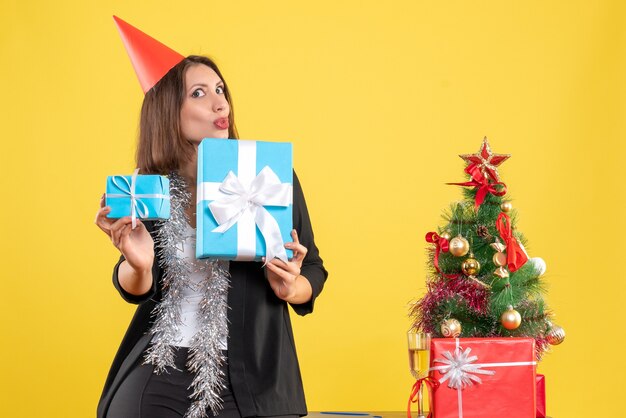 Weihnachtsstimmung mit überraschter positiver schöner Dame mit Weihnachtshut, der Geschenk im Büro auf Gelb hält