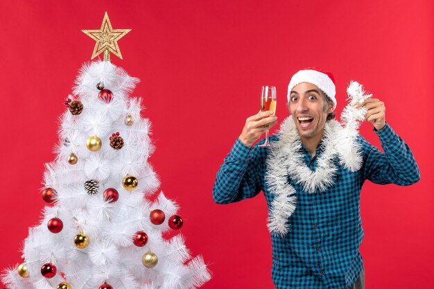 Weihnachtsstimmung mit verrücktem jungem Mann mit Weihnachtsmannhut und einem Glas Wein jubelt sich nahe Weihnachtsbaum