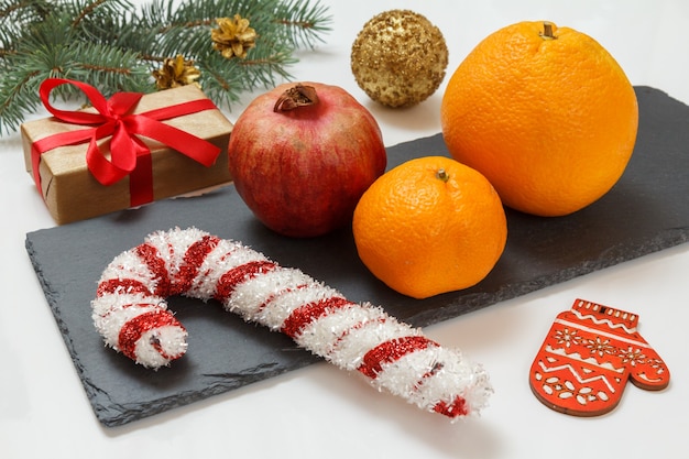 Weihnachtstannenzweig mit zapfen, einer geschenkbox, einer orange, einer mandarine und einem granatapfel auf dem steinbrett und dem weißen hintergrund