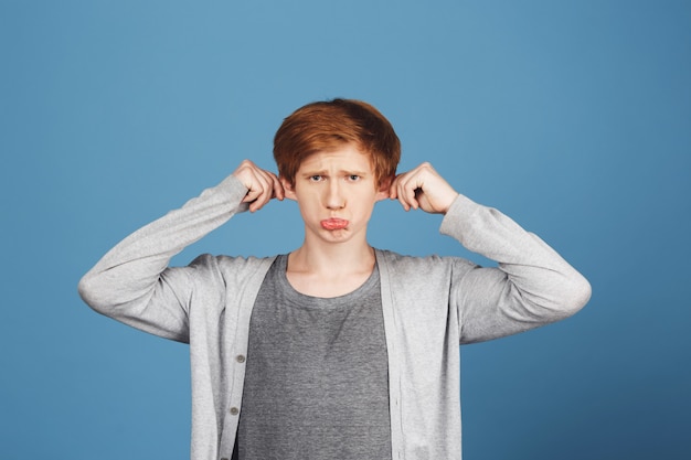 Kostenloses Foto weine nicht, süß. nahaufnahme des jungen gutaussehenden lustigen kerls mit ingwerhaar im lässigen grauen outfit, das ohren mit beiden händen hält, dummen traurigen ausdruck macht und versucht, kleine schwester zum lachen zu bringen.