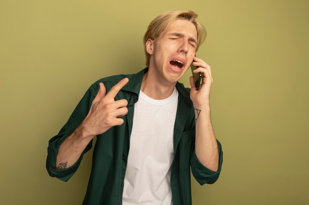 Weinen mit geschlossenen Augen junger blonder Kerl, der grünes T-Shirt trägt, spricht am Telefon