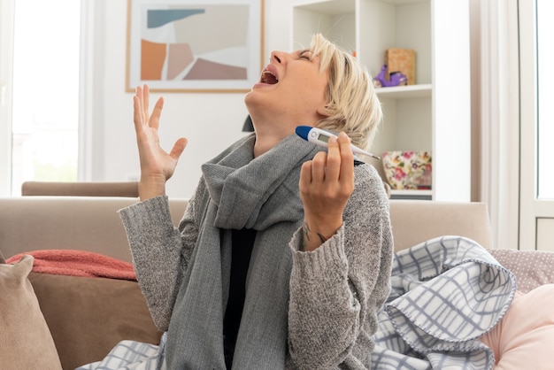 weinende junge kranke slawische frau mit schal um den hals, die ein thermometer hält und auf der couch im wohnzimmer sitzt