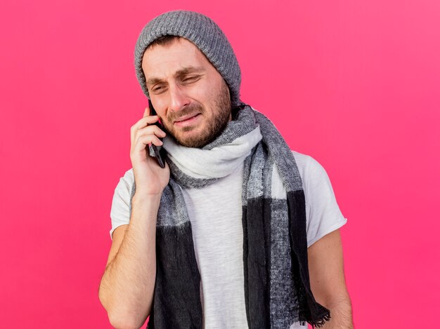 Weinender junger kranker Mann, der Wintermütze mit Schal trägt, spricht am Telefon lokalisiert auf rosa Hintergrund