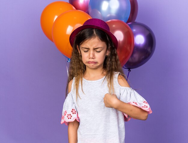 weinendes kleines kaukasisches Mädchen mit violettem Partyhut, das vor Heliumballons steht, isoliert auf lila Wand mit Kopierraum