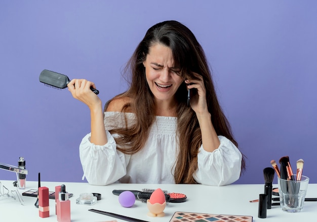 Weinendes schönes Mädchen sitzt am Tisch mit Make-up-Werkzeugen hält Haarkamm, der am Telefon spricht, lokalisiert auf lila Wand