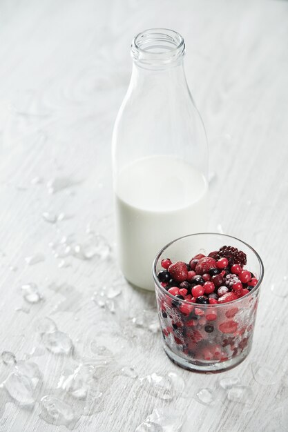 Weinleseflasche mit Milch nahe Glas mit gefrorenen Beeren bereit für Cocktailherstellung lokalisiert auf weißem Tisch mit geschmolzenem Eis