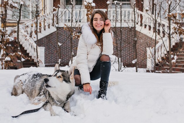 Weiße Frau mit erstaunlichem Lächeln, das mit ihrem Hund während des Winterspaziergangs im Hof aufwirft. Außenfoto der fröhlichen Dame trägt zerrissene Jeanshosen, die auf dem Schnee mit faulem Husky sitzen.