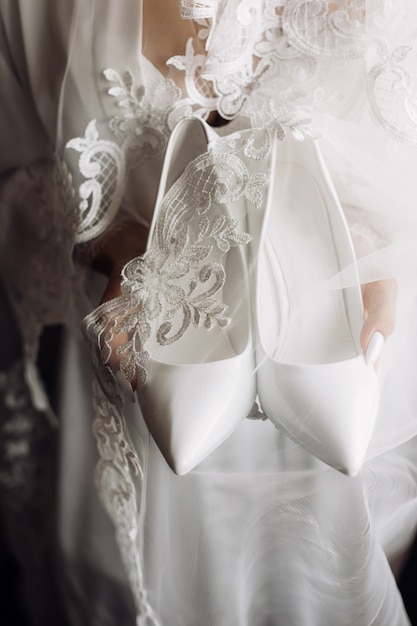 Weiße Hochzeitszeremonienschuhe in den Händen der Braut gekleidet in Seidennachtwäsche mit Spitze