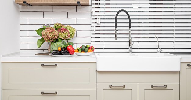 Weiße Küchenschränke mit Metallgriffen an den Türen in der Nähe des Waschbeckens mit einem Blumenstrauß und einem Gemüseteller.