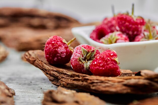 Weiße Schüssel mit frischen Himbeeren mit Holzstücken auf grauer Oberfläche. Nahansicht.