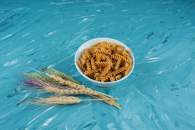 Weiße Schüssel trockene Fusilli-Nudeln mit Weizenähren auf blauem Hintergrund.