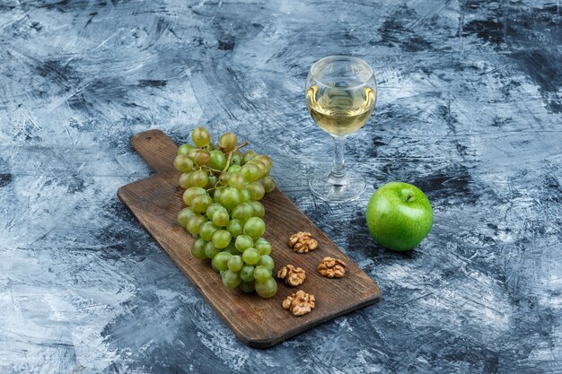 Weiße Trauben der hohen Winkelansicht, Walnüsse auf Schneidebrett mit Glas Whisky, grüner Apfel auf dunkelblauem Marmorhintergrund. horizontal