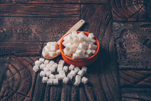 Kostenloses Foto weiße zuckerwürfel in einer orangefarbenen schüssel mit hoher winkelansicht des löffels auf einem dunklen holztisch