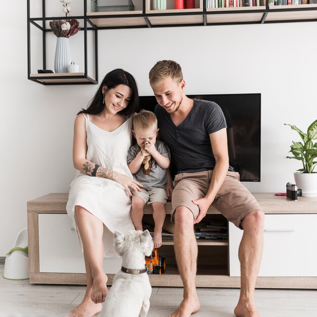 Weißer Hund, der die lächelnden jungen Paare sitzen mit ihrem Sohn vor Fernsehen betrachtet