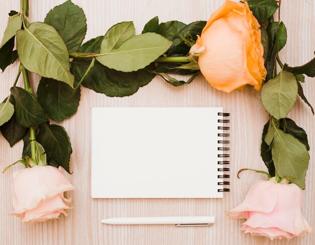 Weißer Stift und leerer gewundener Notizblock umgeben mit Rosen über hölzernem Hintergrund