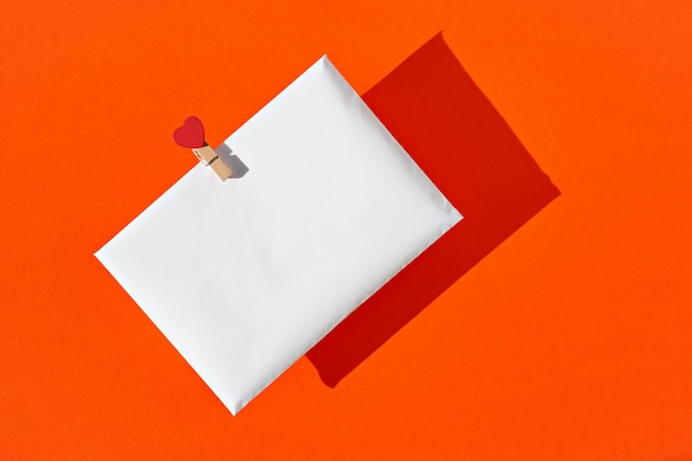 Weißer Umschlag mit rotem Herz auf einer Wäscheklammer auf orangefarbenem Hintergrund. Liebesbrief oder Glückwünsche zum Valentinstag, Muttertag, Frauentag. Sicht von oben