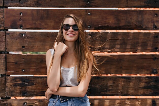 Weißes Mädchen, das gute Gefühle auf Holzwand ausdrückt. Porträt der prächtigen blassen jungen Frau trägt dunkle Sonnenbrille.