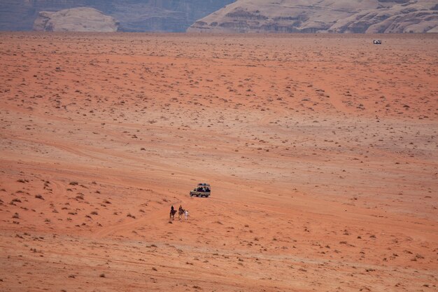 Weitwinkelaufnahme von zwei Personen auf Kamelen, die sich einem Auto in einer Wüste nähern