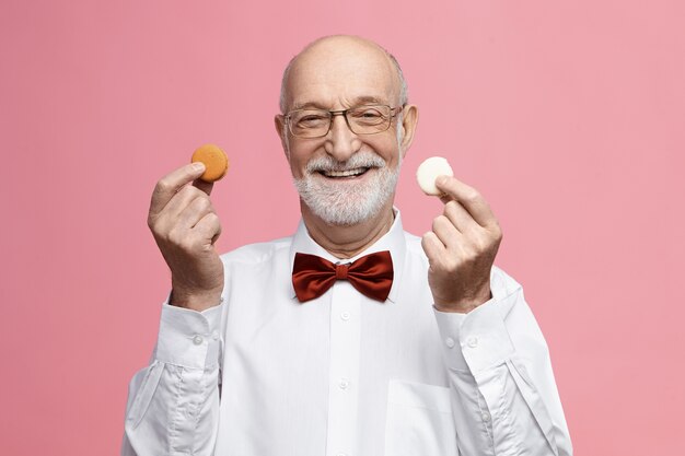 Welches magst du? isoliertes bild des fröhlichen energischen älteren mannes im ruhestand, der brille und fliege trägt, breit lächelt, bunte macarons in jeder hand hält und ihnen anbietet, einige zu haben