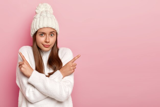 Welches zu wählen. Zögernde natürliche europäische Frau zeigt seitwärts, verschränkt die Arme über der Brust, schlägt zwei Varianten vor, trifft Entscheidung, trägt warmweißen Pullover und Hut, Leerzeichen auf rosa Hintergrund