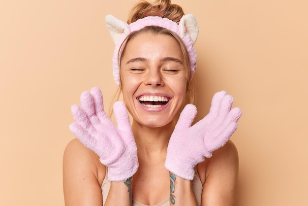 Wellness- und duschkonzept für die körperpflege. positive junge frau lächelt gerne und benutzt badehandschuhe zum waschen und entfernen abgestorbener hautzellen, trägt stirnband-posen vor beigem hintergrund.