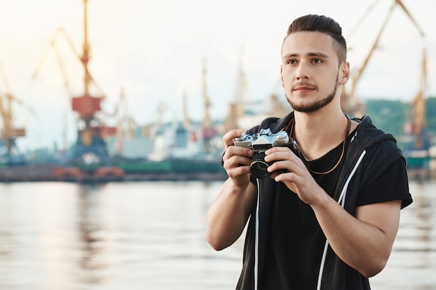 Wenn Hobby zur geliebten Arbeit wird. Porträt des verträumten kreativen jungen Kerls mit Bart, der Kamera hält und mit nachdenklich erfreutem Ausdruck beiseite schaut und Fotos von Hafen und Meer beim Gehen macht