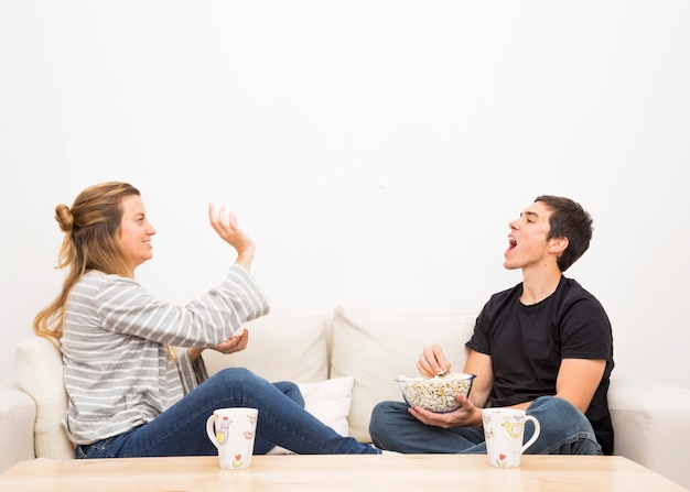 Werfendes Popcorn der Frau im Mund des Mannes, der auf Sofa sitzt