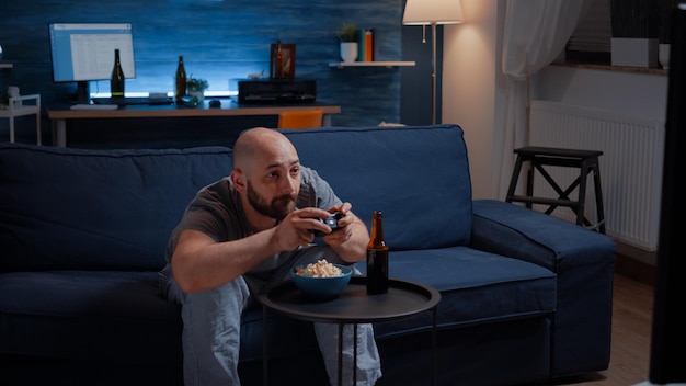 Wettbewerbsfähiger entschlossener Mann, der nachts im Wohnzimmer Videospiele spielt, konzentriert sich auf das Essen von Popcorn und genießt die Freizeit. Beteiligter Spieler, der auf der Couch sitzt und Videospiele mit Joystick, Wireless-Controller spielt