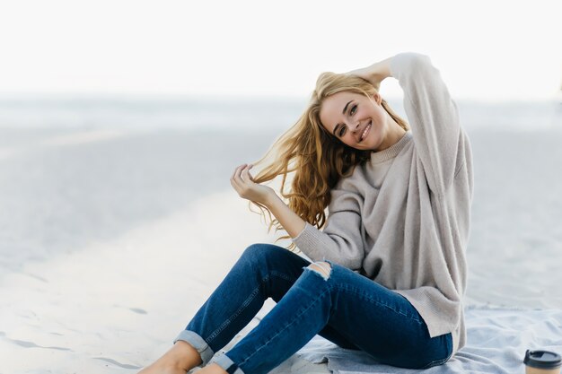 Wicklerin, die am Herbststrand emotional aufwirft. Außenporträt der hübschen lockigen Frau in den Jeans, die im Sand sitzen.