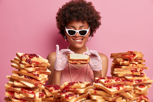 Wie lecker! Erfreute afroamerikanische Dame in Modekleidung, Spitzenhandschuhen, trendiger Sonnenbrille, auf Bankett, posiert in der Nähe von Brottoast, isoliert auf rosa Wand, hält appetitliches Sandwich