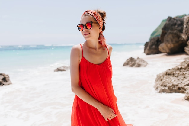 Winsome frau im roten kleid, das am strand am wochenendmorgen aufwirft. sorgloses kaukasisches mädchen in den trendigen sommeraccessoires, die im strand stehen