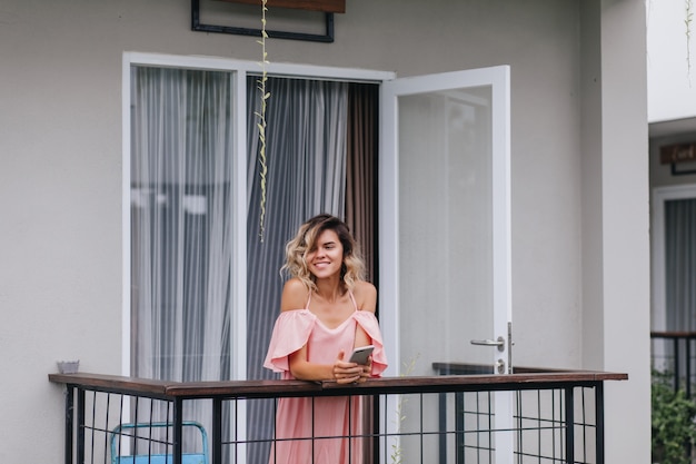 Winsome gebräuntes Mädchen mit Telefon in den Händen lächelnd und weg schauend. Fröhliche junge Dame im rosa Outfit, die am Hotelbalkon steht.