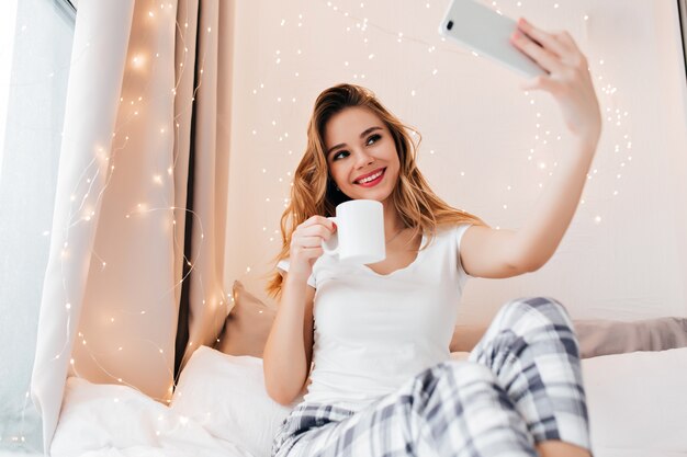 Winsome Mädchen, das morgens Tee trinkt und sich selbst fotografiert. Lächelnde herrliche Frau, die Kaffee im Schlafzimmer genießt und Selfie macht.