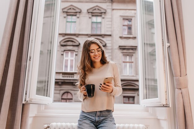 Winsome Mädchen in der Freizeitkleidung, die Zeit zu Hause verbringt und auf Stadthintergrund mit Tasse Tee aufwirft