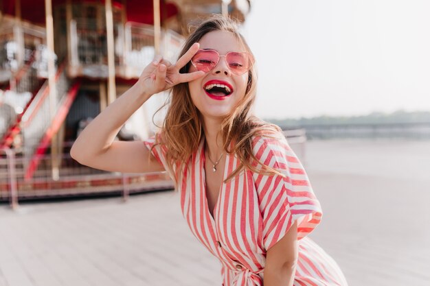 Winsome weibliches Modell im gestreiften Weinlesetanz, der im Vergnügungspark tanzt. Außenporträt der glückseligen blonden Frau in der Sonnenbrille, die nahe Karussell mit Friedenszeichen steht.