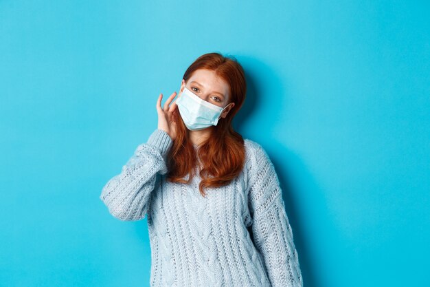 Winter, Covid-19 und Konzept der sozialen Distanzierung. Süßes rothaariges Teenager-Mädchen, das Gesichtsmaske trägt und die Haarsträhne hinter dem Ohr steckt, in die Kamera starrt und auf blauem Hintergrund steht