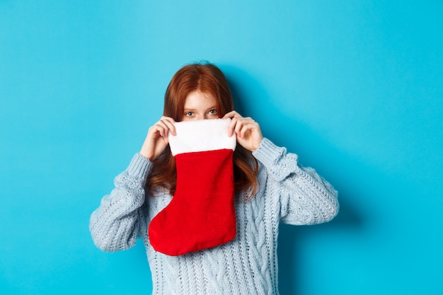 Winterferien und Geschenkkonzept. Lustiges rothaariges Mädchen, das in den Weihnachtsstrumpf schaut und mit den Augen lächelt und vor blauem Hintergrund steht