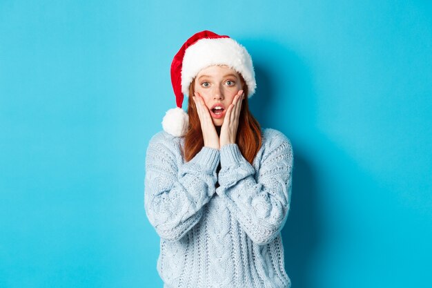 Winterferien und Heiligabend-Konzept. Überraschtes rothaariges Mädchen in Weihnachtsmütze, das ungläubig in die Kamera starrt, offener Mund erstaunt, über blauem Hintergrund stehend