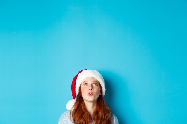 Winterferien und Heiligabend-Konzept. Kopf eines hübschen rothaarigen Mädchens in Weihnachtsmütze, erscheint von unten und schaut auf das Logo beeindruckt, sieht Promo-Angebot, blauer Hintergrund