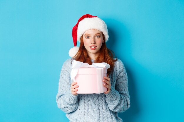 Winterferien und Heiligabend-Konzept. Nettes rothaariges Mädchen, das Sankt-Hut trägt, Geschenk des neuen Jahres hält und Kamera betrachtet, stehend vor blauem Hintergrund.