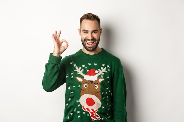 Winterferien und Weihnachten. Zufriedener bärtiger Mann im grünen Pullover, der ein OK-Zeichen zeigt, wie etwas Gutes, das auf weißem Hintergrund steht.