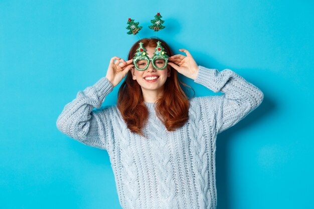 Winterferien und Weihnachtsverkaufskonzept. Schönes rothaariges weibliches Modell, das Neujahr feiert, lustiges Partystirnband und eine Brille trägt, albern lächelt, blauer Hintergrund