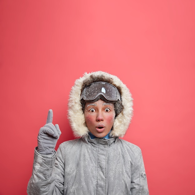 Winterferienkonzept. Die geschockte, gefrorene junge Frau hält den Mund weit offen und zeigt nach oben, betäubt von sehr niedrigen Temperaturen und starkem Schneefall, geht im Dezember in Oberbekleidung Skifahren