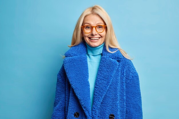 Wintermode-Konzept. Glücklich lächelnde blonde Frau des Mittelalters trägt optische Brille und blauen Mantel.