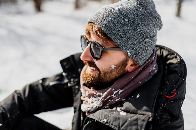 Winterporträt des Hipster-Mannes mit Bart im grauen Hut, der im sonnigen Park mit Schneeflocken auf Kleidern entspannt