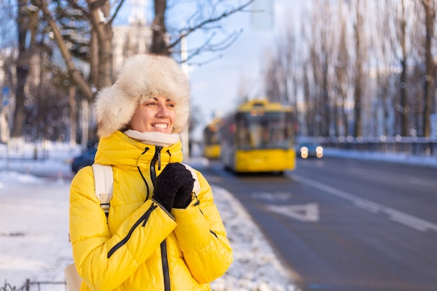 Winterporträt einer glücklichen Frau in einer warmen gelben Jacke und einem sibirischen russischen Hut, die auf einen Bus auf einer verschneiten Stadtstraße warten