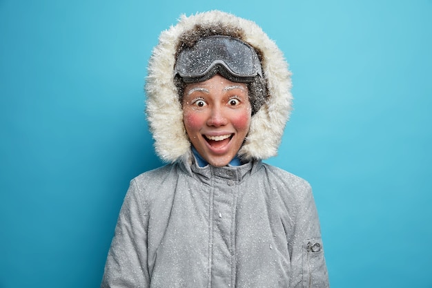 Wintersport- und Freizeitkonzept. Glückliche fröhliche aktive Frau mit dem roten gefrorenen Gesicht auf Schneesturm, der nach dem Skifahren zufrieden ist, hat Spaß während des kalten Tages auf der Wanderreise gekleidet in der grauen Jacke.