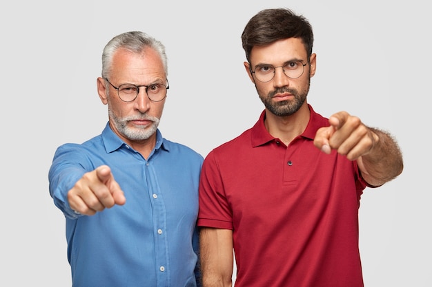 Wir wählen genau Sie! Zuversichtlich ernst zeigen zwei Männer mit Zeigefingern, drücken ihre Wahl aus, tragen helle Kleidung, isoliert über weißer Wand. Älterer Mann mit erwachsenem Sohn innen