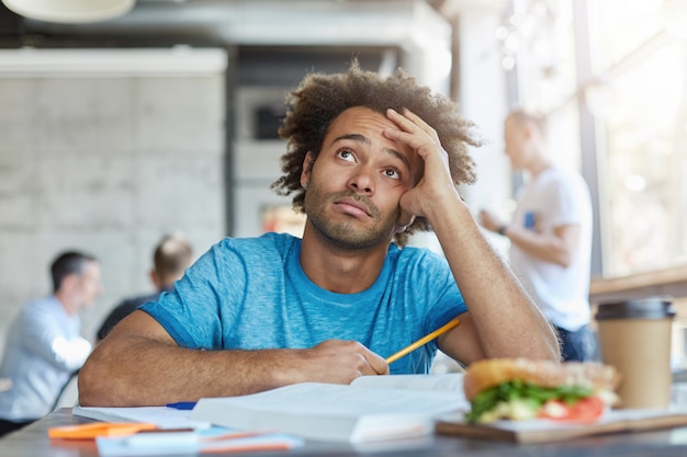 Wissen und Bildung. Unglücklicher afroamerikanischer Universitätsstudent, der blaues T-Shirt trägt, das mit fragwürdigem frustriertem Ausdruck aufblickt und sich müde fühlt, während er an Hausaufgabe im Café arbeitet