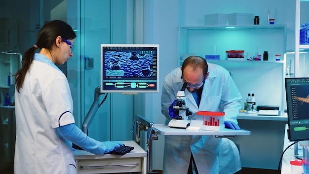 Wissenschaftlerforscher mit Mikroskop im Labor, während Krankenschwester Notizen über Computer macht, die Überstunden machen